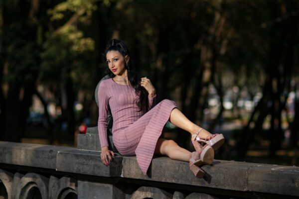 Woman in pink dress and heels sits elegantly on a stone bench in a park setting.